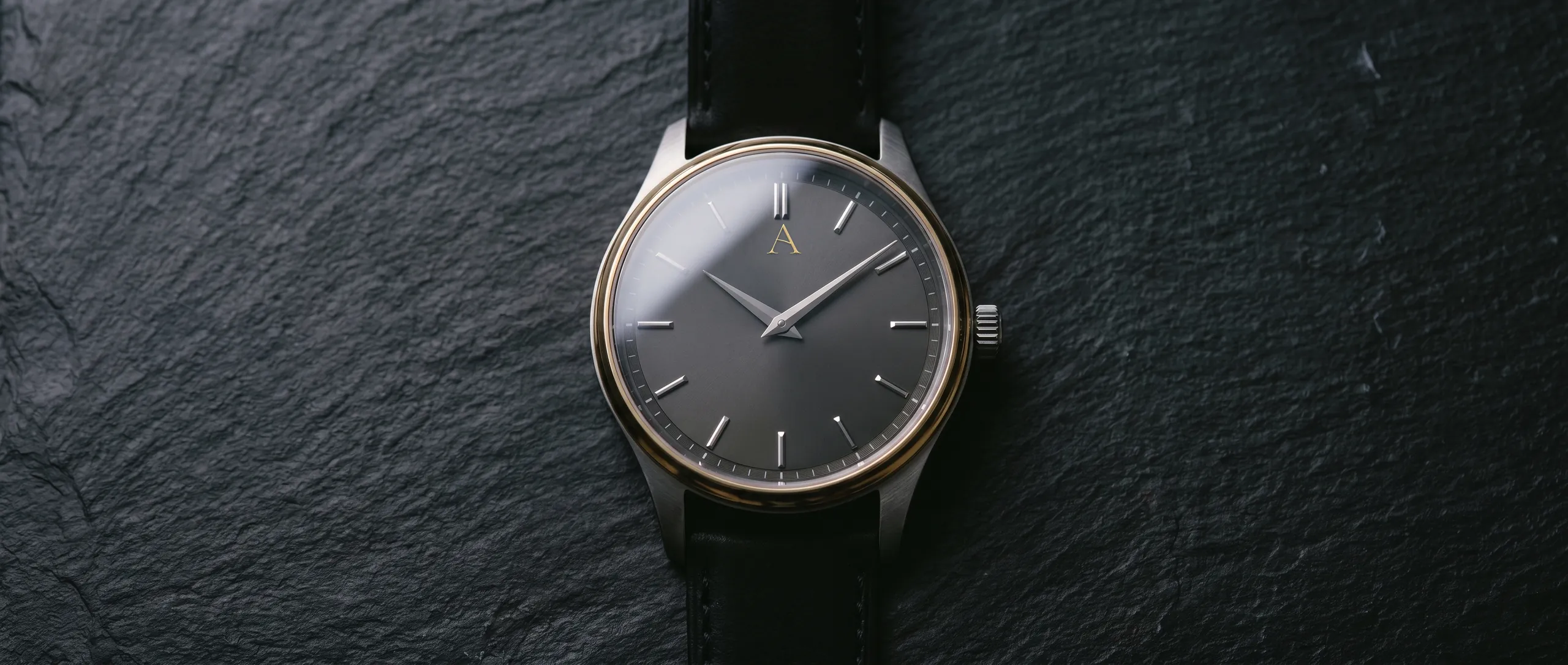 Montre acier et or au cadran gris ardoise, le A en applique, sur pierre sombre.